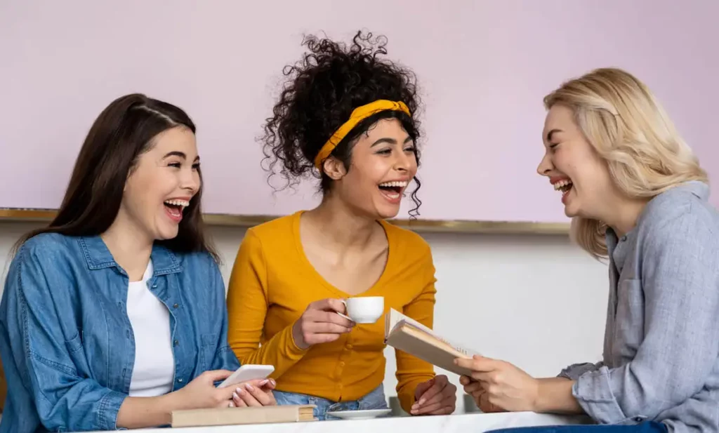 Três amigas se divertindo em um café, rindo e compartilhando momentos, segurando livros e um copo de café, em um ambiente descontraído.