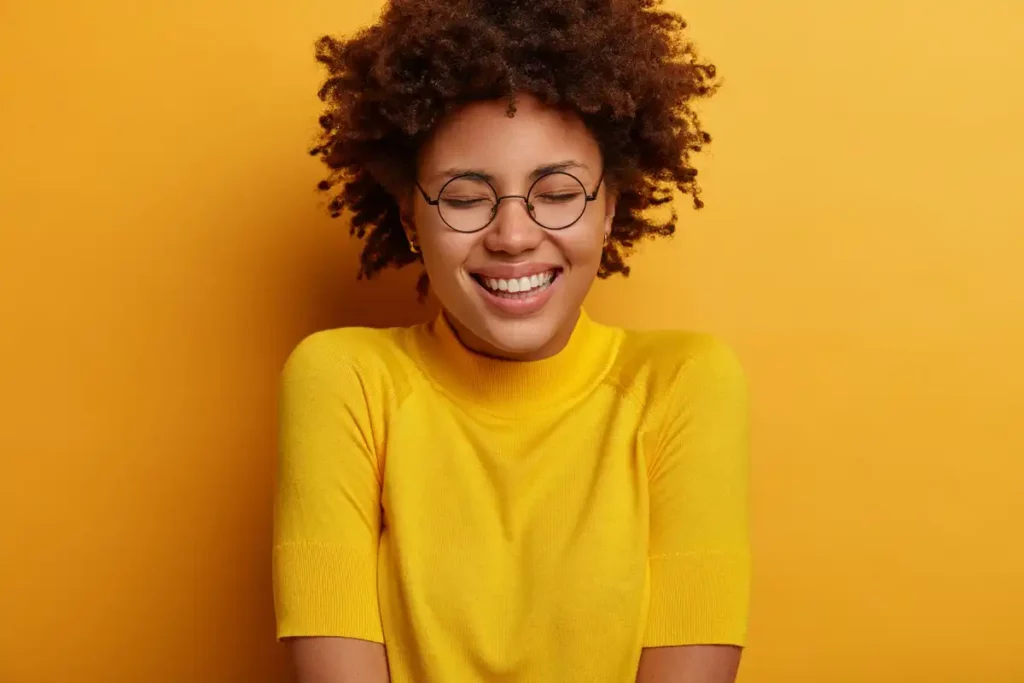 Mulher sorridente com cabelo cacheado e óculos, vestindo uma blusa amarela contra um fundo amarelo.