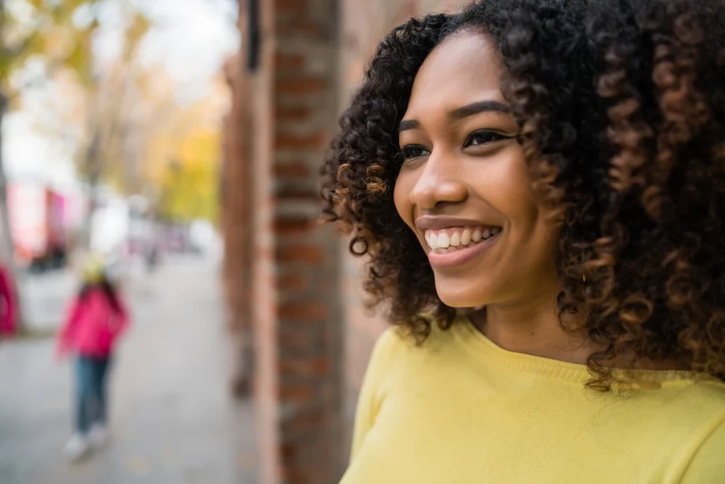 Mulher sorridente com cabelo cacheado, usando uma blusa amarela, ao ar livre em uma rua com árvores e pessoas ao fundo.