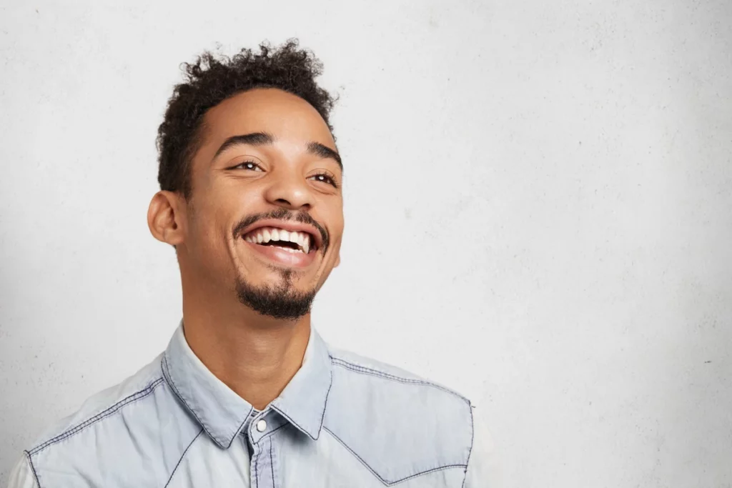 Homem sorridente com cabelo cacheado, usando uma camisa jeans, em um fundo claro, expressando felicidade e alegria.